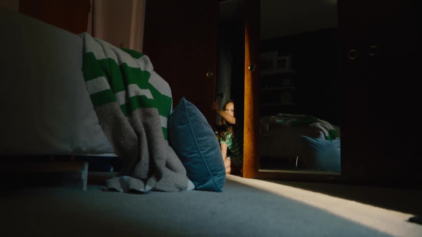 A young girl hides in a dimly lit closet during a game of hide and seek