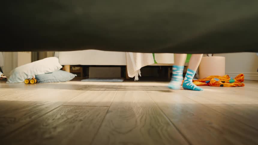 A child wearing fun socks walks away from a bed viewed from a low angle beneath the furniture