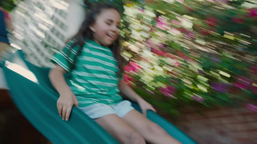 A girl joyfully slides down outdoor slide with boy in background