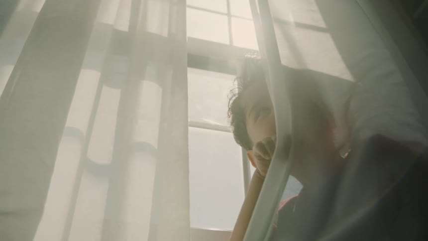 Boy peeks out playfully from behind curtain in soft daylight