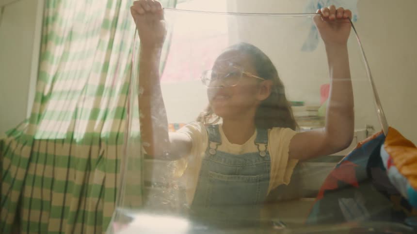 Joyful girl playfully hides behind glass under a blanket fort as sunlight streams in