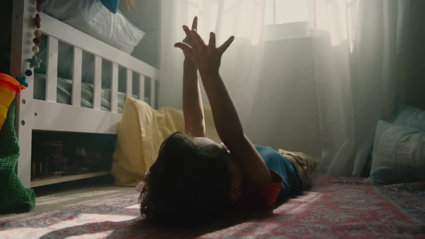 Child lies on bedroom floor playfully counting fingers in sunlight