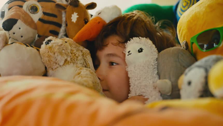 A young boy peeks out joyfully from a pile of stuffed animals before hiding his face again
