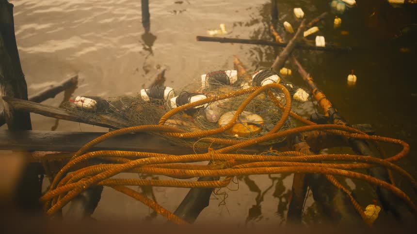 A tangled fishing net with floats rests on weathered wooden planks above murky water littered with debris
