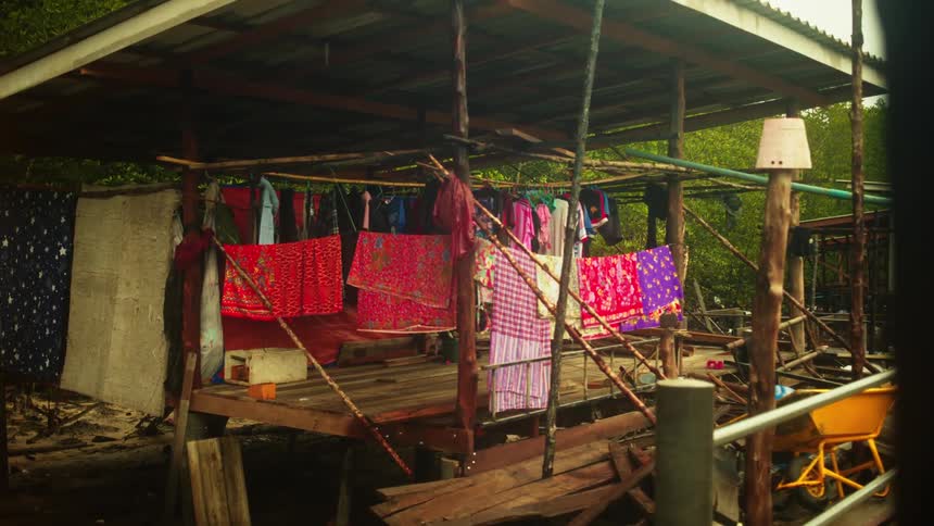 A rustic wooden stilt house with a corrugated metal roof stands amidst lush green foliage adorned with colorful clothes hanging to dry on a clothesline