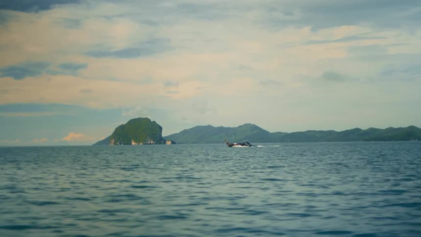 A boat moves across rippling turquoise water along a horizon dotted with verdant rocky islands under a partly cloudy sky