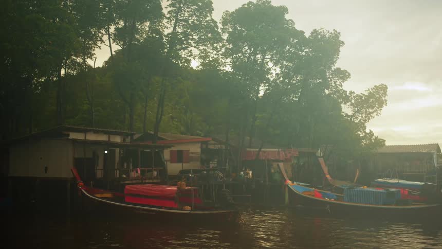Stilted shacks and docked boats line a murky riverbank under an overcast sky