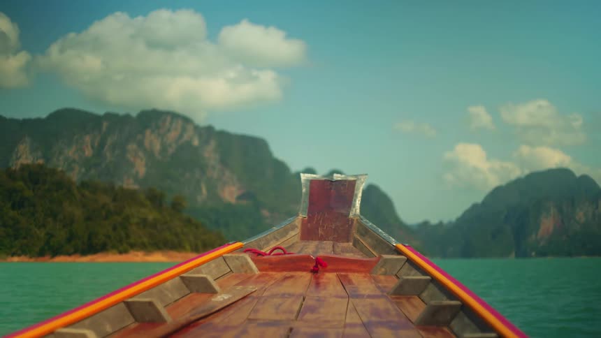 A wooden longtail boat navigates turquoise waters toward towering jungleclad cliffs under a partly cloudy sky
