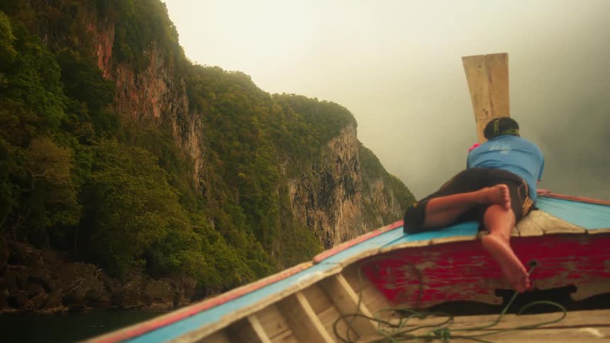 A man is laying on the boat traveling past the mountains