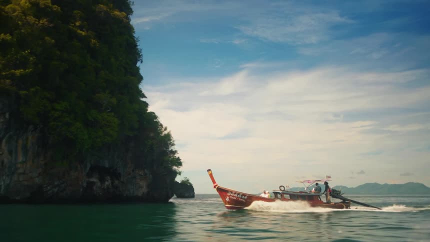 Thai boat cruising through crystal clear waters along a forested coastline