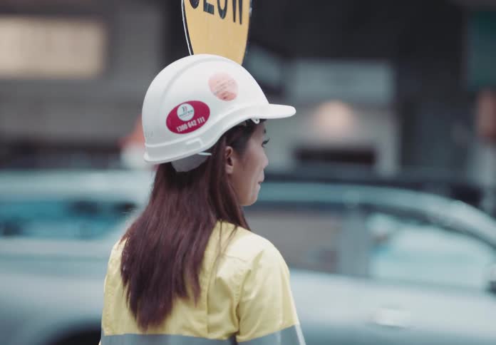 A construction worker wearing a hard hat and a yellow jacket is seen from behind standing on a city street holding a slow sign
