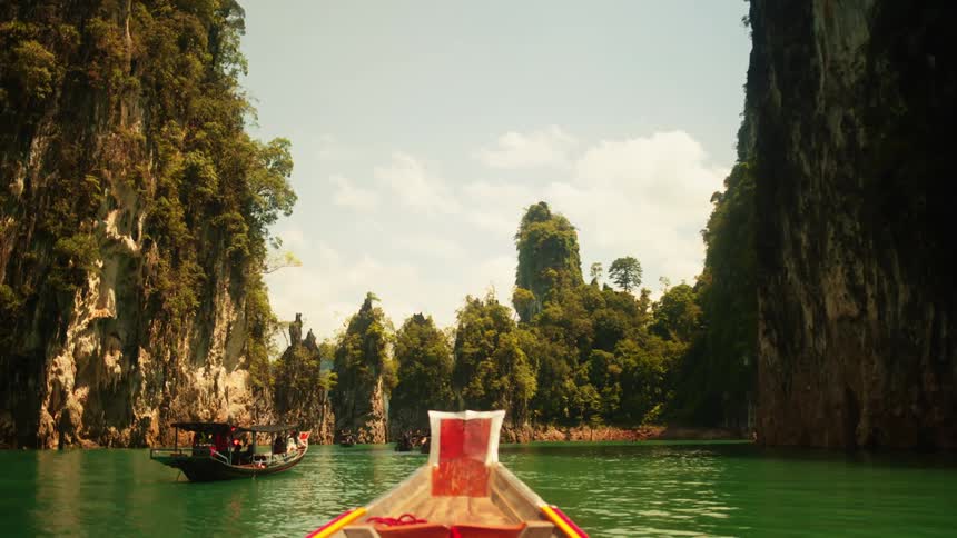 One boat passes another floating on green water between jungle covered cliffs on a cloudy day