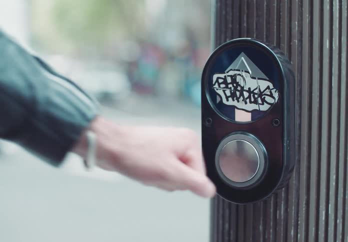 A womans fist presses a pedestrian call button at the side of a busy road as vehicles pass by at midday