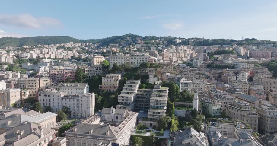 An aerial shot slowly advances over a densely built cityscape towards a terraced complex of buildings nestled amongst lush trees backed by a verdant building covered hillside under a clear blue sky