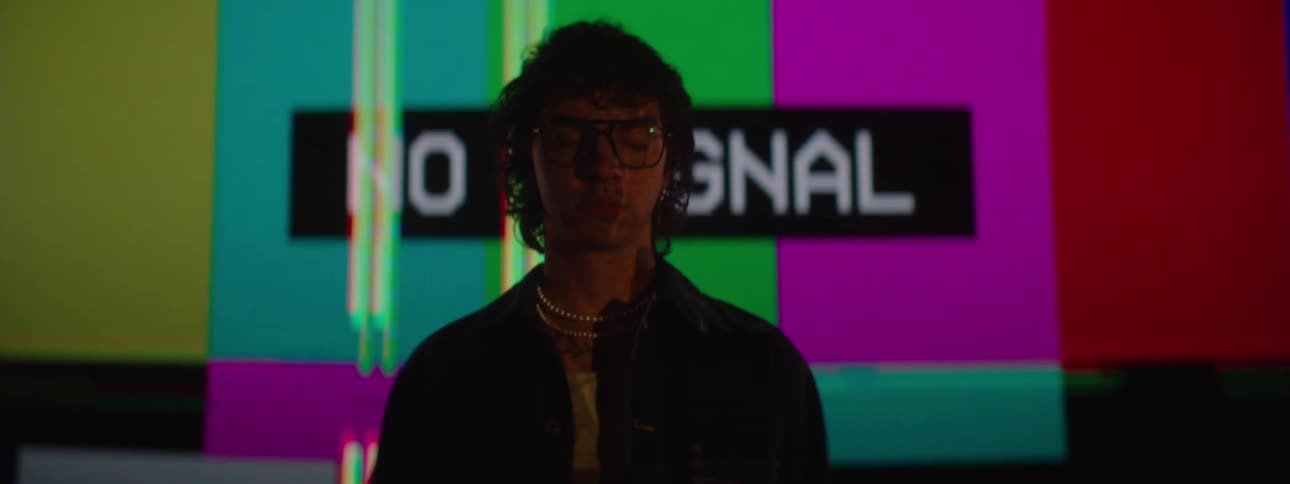 A young man wearing glasses stands in front of a projection screen displaying a no signal message followed by distorted colors that shine behind and through him