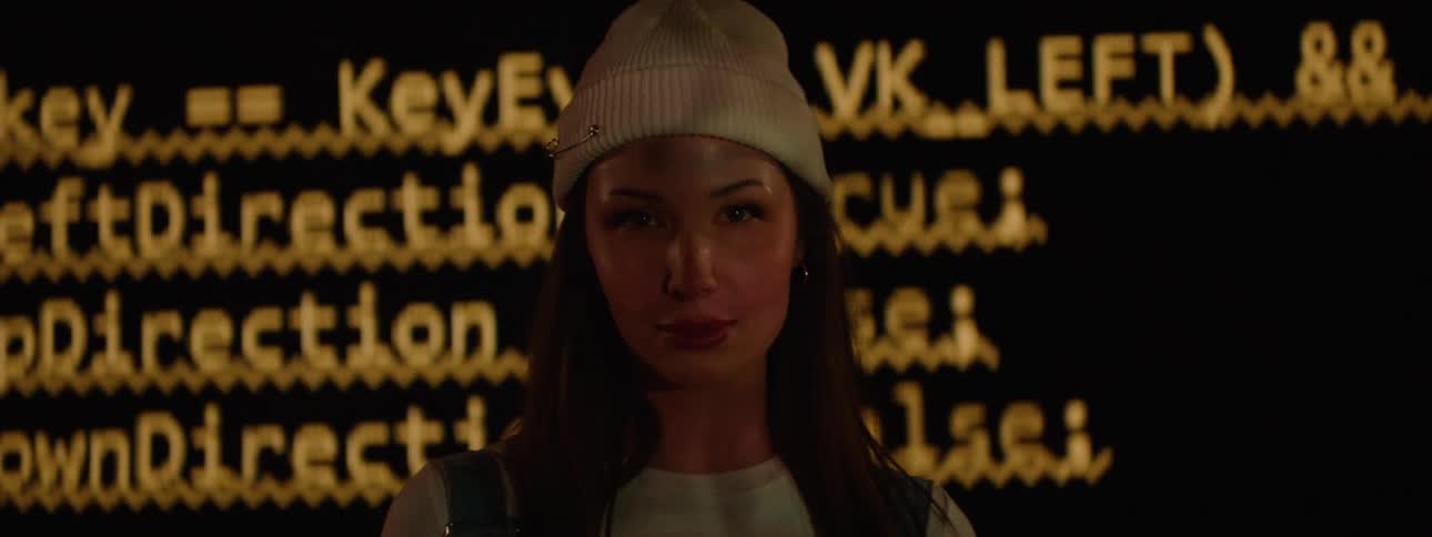 A woman with a slight smile walking forward with a large screen behind her showing text and programming code