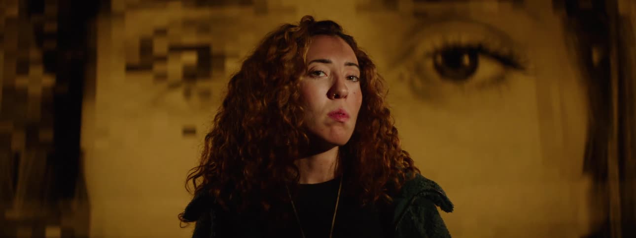 A woman with curly hair wearing a dark jacket and necklace stands in front of a large slightly out of focus mural of a persons eyes in a warm yellow light