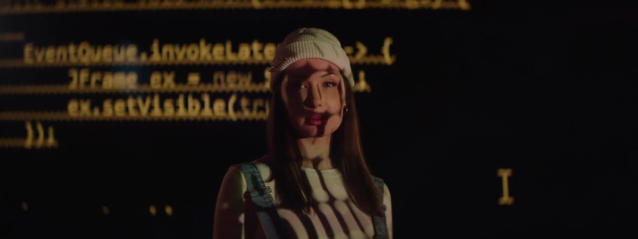 A young woman stands in front of a projector screen displaying a series of letters that appear both behind her and projected onto her body she is wearing a hat and the room is dark