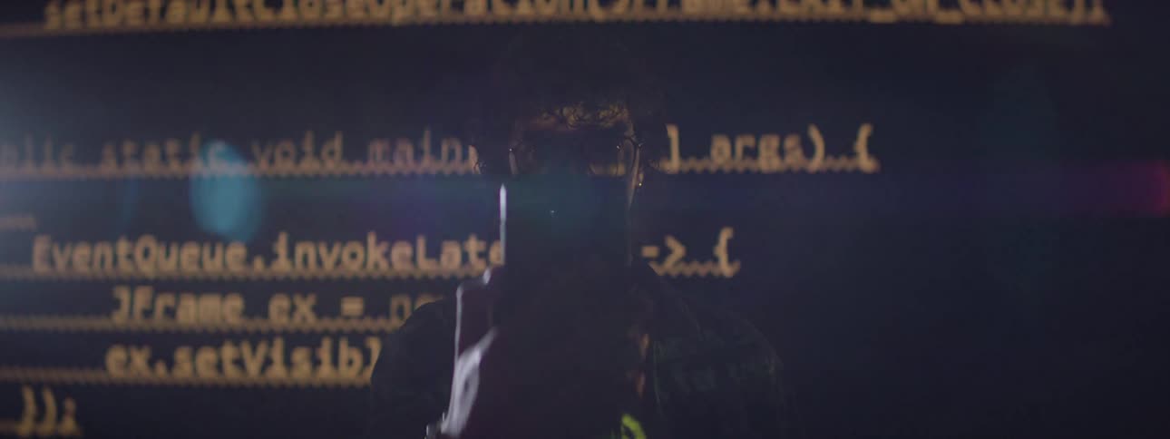 A person wearing glasses is backlit by scrolling computer code while holding a phone in front of their face