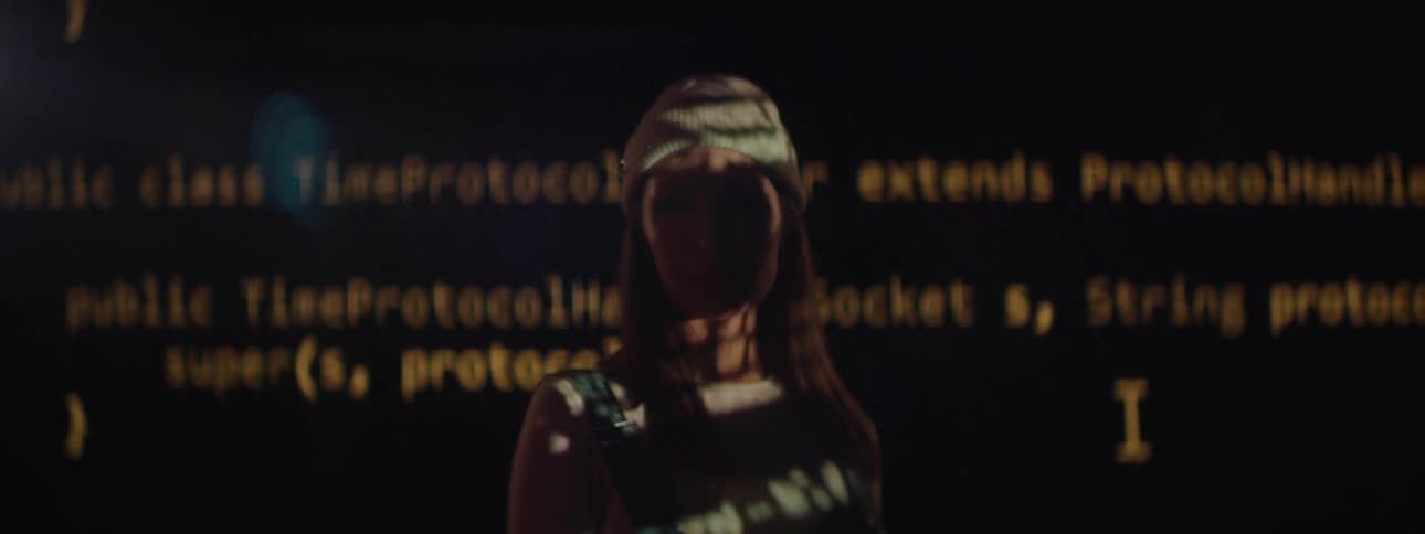 A large screen with code on it and a woman standing in front of it staring straight ahead