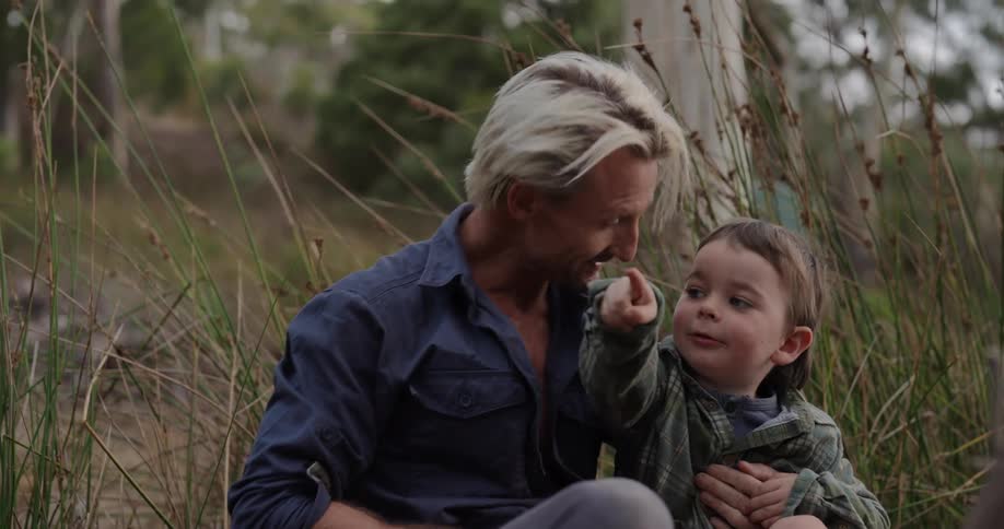 A father and son sit on a grassland engaging with each other excitedly under the midday sun