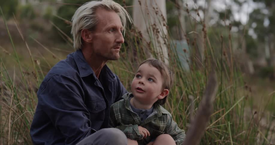 A man sitting on the ground with a child near a tree and tall grass pointing forward