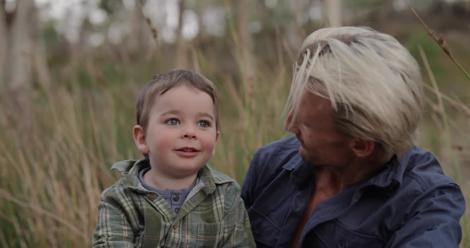 A man with blond hair holds a child with bright blue eyes in his lap and the child points up towards something in the sky