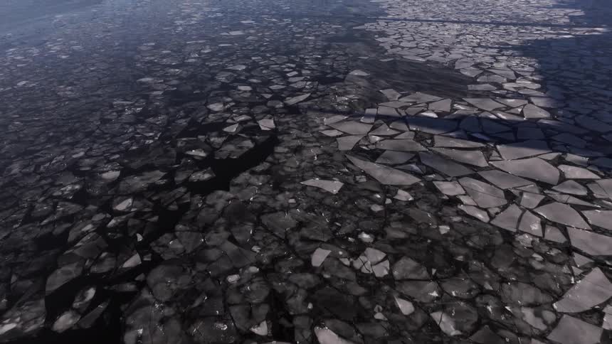 An aerial shot reveals a body of water partially covered in fragmented ice floes gradually transitioning to a clear expanse with a cityscape skyline visible in the distance under a bright sky