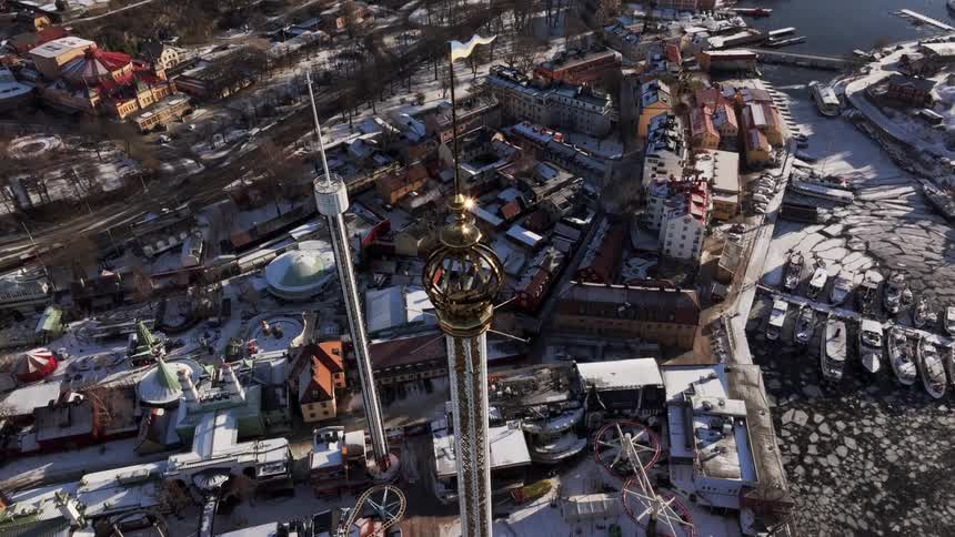 An ornate crown of a tower with a flag stands above a snow dusted amusement park adjacent to a frozen waterfront