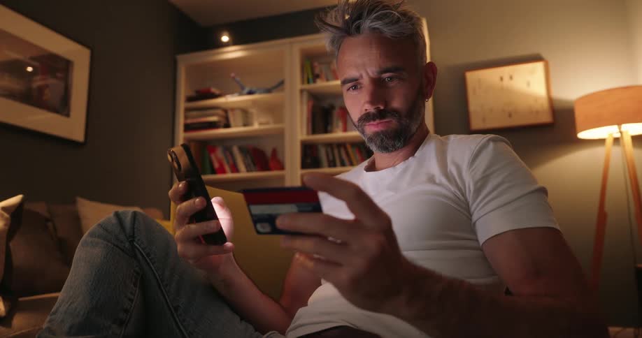 A man with salt and pepper hair and a beard is sitting on a couch holding a credit card in one hand and looking at his phone in the other