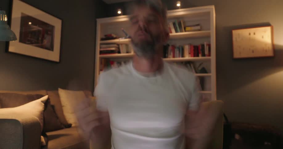 A man with grey hair and a beard sits on a couch in a dimly lit room holding a smartphone and a credit card seemingly focused on an online transaction