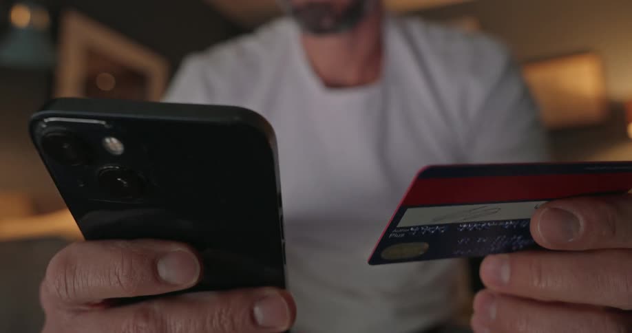A man sitting indoors holds a phone in one hand and a credit card in the other flipping the card around while copying the number onto the phone