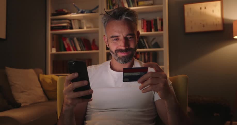 A smiling man with gray hair and a beard sits comfortably looking at his smartphone while holding a credit card suggesting he is likely engaged in online shopping