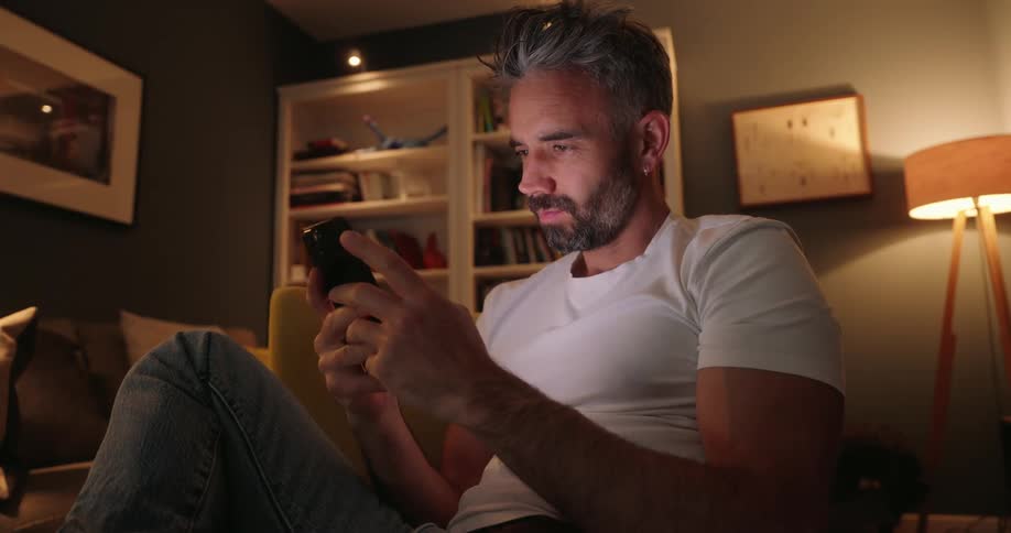In a dimly lit room a man with grey hair and a beard sits on a couch and intently uses his smartphone