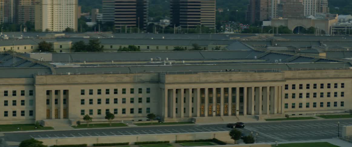 A large city building with taller buildings behind it and a parking lot