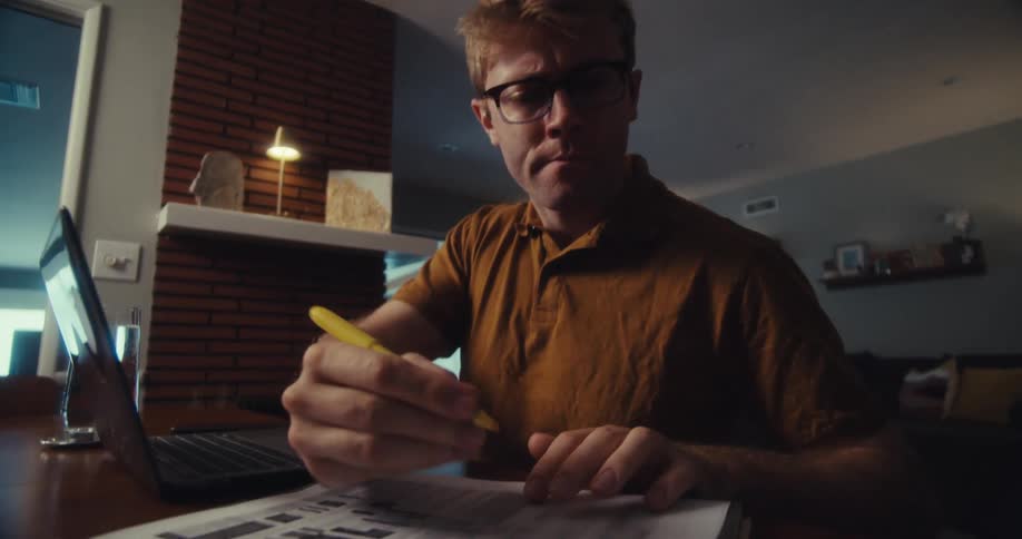 Man sitting at a table at home consults a computer before highlighting information in a document