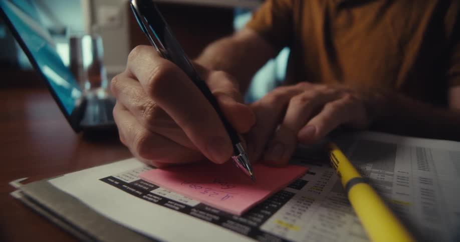 A man writes financial notes on a pink sticky note beside a printed spreadsheet and open laptop