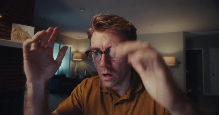 A young man sitting behind his computer with glasses on suddenly changes his expression and places both hands on his cheeks in disbelief while in his room