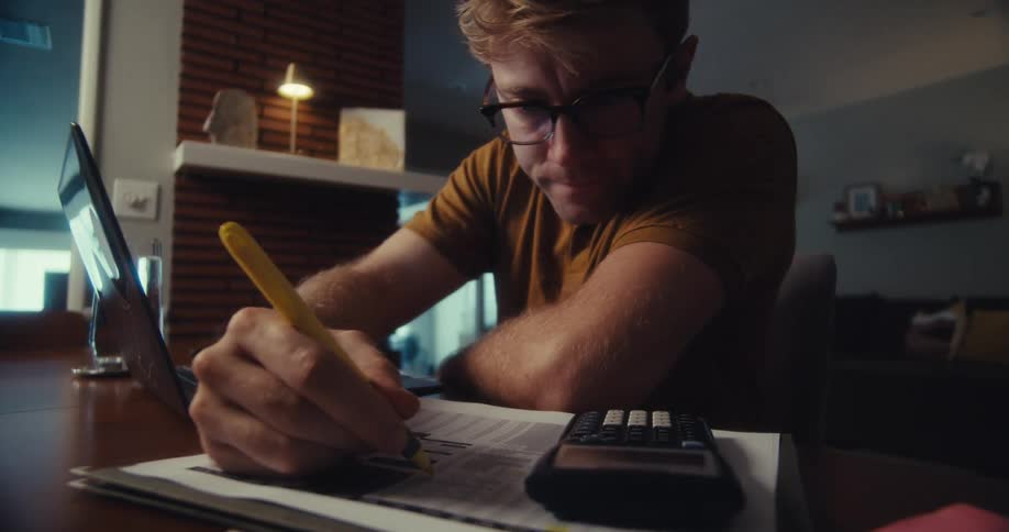 A man wearing glasses works on his laptop indoors then picks up a pen and writes in a notebook beside him