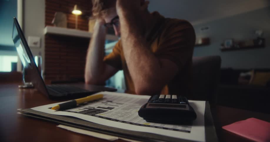 A frustrated man wearing glasses sits at a table with a laptop calculator and financial documents appearing stressed as he tackles his finances