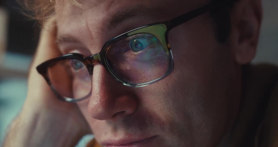 A man wearing glasses reacts emotionally while looking at his laptop screen in a softly lit indoor space