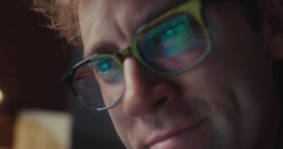 A man stares intensely at a laptop screen reflected in his glasses suggesting deep focus or concern