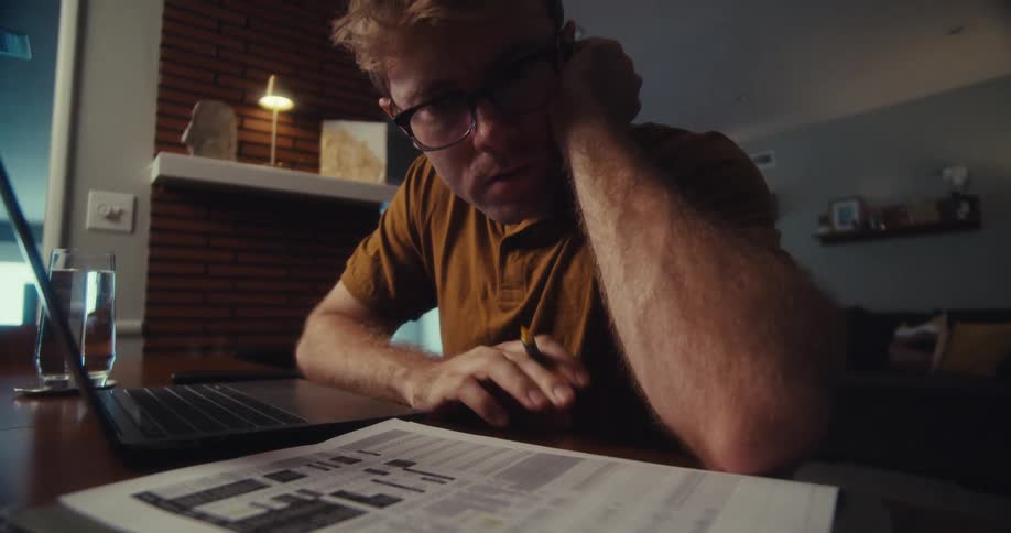 Man with a pencil sitting at a table with an open laptop and analyzing a document