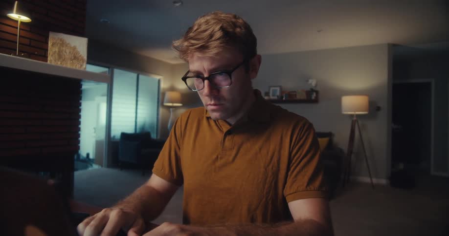 A man types on his laptop while appearing visibly worried in a dimly lit home setting