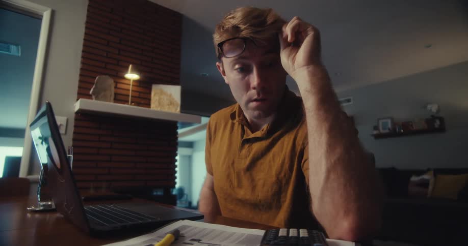 A worried man calculates finances while looking at his laptop and pressing a calculator