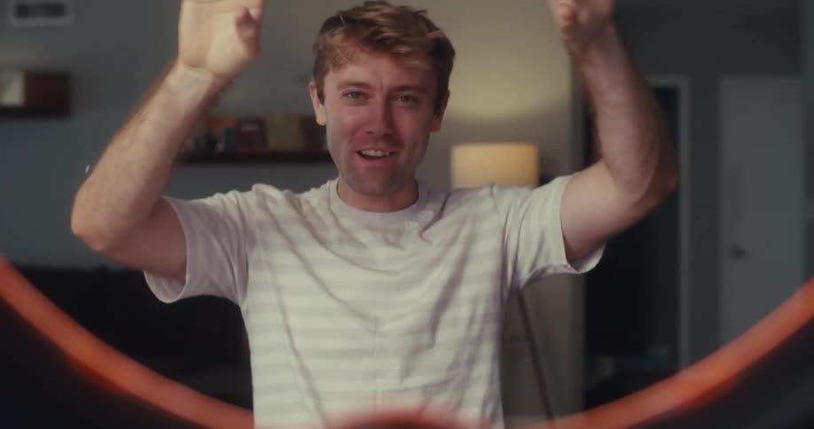 A cheerful man is vlogging in a cozy indoor setting enthusiastically pointing downward while speaking to the camera