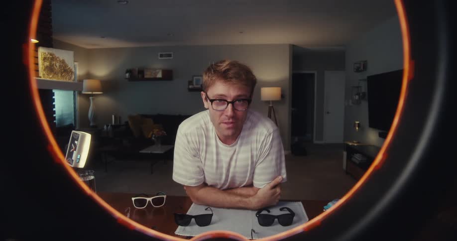 A man sitting behind a table wearing glasses with several other glasses laid out in front of him talks and samples two of them to the camera makes hand gestures and then walks out of the sitting room