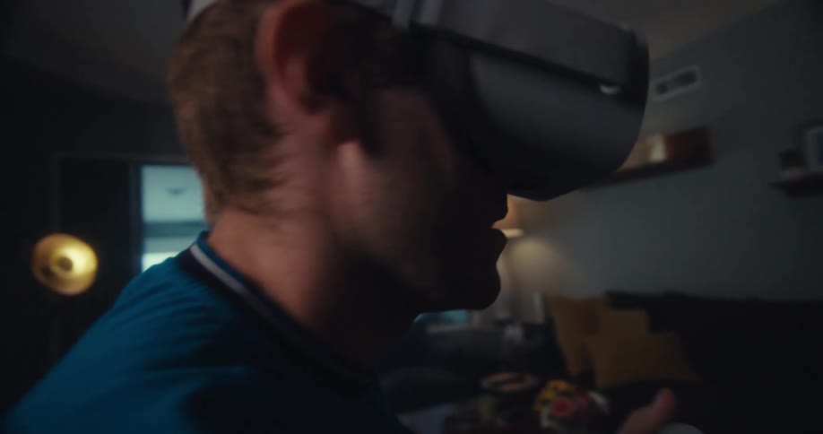 A man wearing a vr headset sits in a living room fully immersed in virtual reality