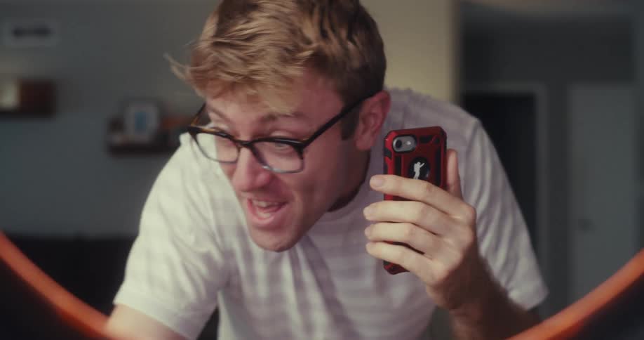 A man enthusiastically films himself showcasing sunglasses using his phone in a casual home setting