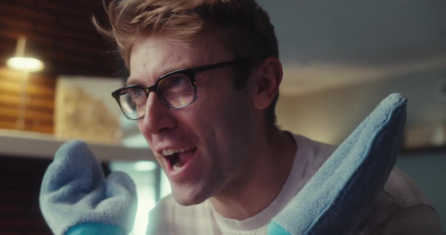 Blonde man wearing oven mitts while standing in living room talking